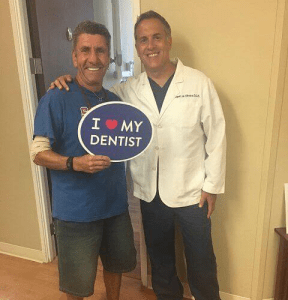 Dr. August de Oliveira with a male patient holding I love my dentist sign beside a man
